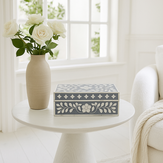 Decorative box with floral pattern on a white table next to a vase with white flowers, in a bright room.