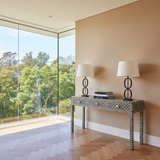 Two table lamps on a decorative console table with large windows showing greenery outside.
