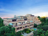Large multi-story building with balconies and a pool, surrounded by trees under a clear blue sky.