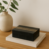 Black decorative box with gold accents on a wooden surface next to a beige vase.