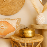 Decorative setting with a gold box on a wicker table, pillows, and a vase with dried plants.