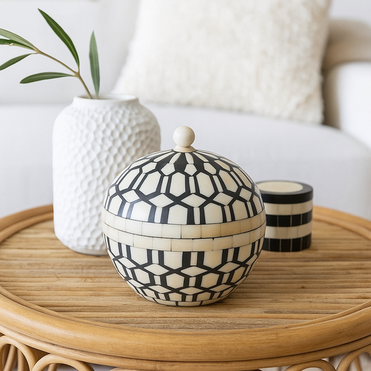 Decorative black and white patterned box on a wooden table with a textured vase and candle in the background.