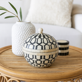 Decorative black and white patterned box on a wooden table with a textured vase and candle in the background.