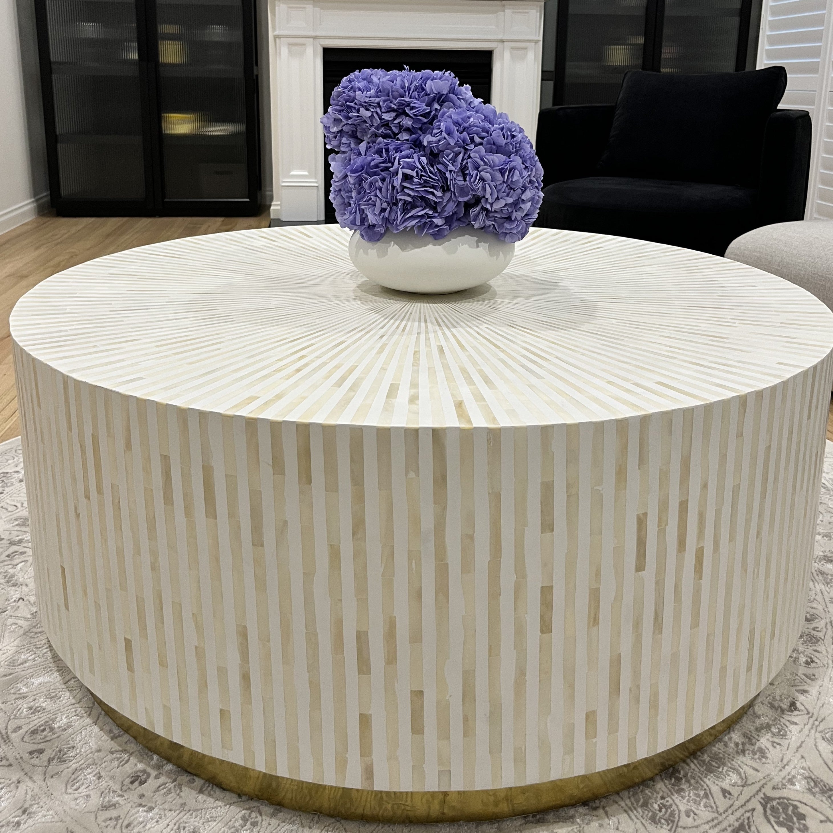 Round coffee table with a decorative purple flower arrangement in a white vase, set against a living room background.