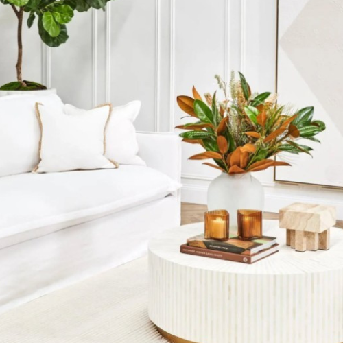 Modern living room with a white sofa, decorative plant, and coffee table with candles and books.