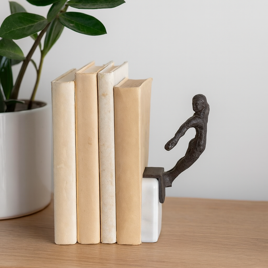 Decorative bookend shaped like a person holding books on a wooden surface with a plant in the background.