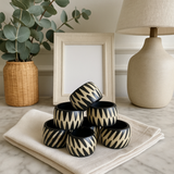 Stack of black and gold patterned containers on a marble surface with a lamp and plant in the background.