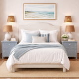 Neatly made bed with white bedding and blue throw, flanked by two nightstands with lamps and a beach-themed painting on the wall.