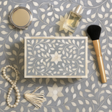 Makeup items including a compact mirror, perfume bottle, brush, and decorative box on a patterned surface.