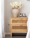 Wooden nightstand with wicker design, vase of flowers, and candles against a neutral wall.
