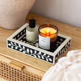 Candle and bottle on a decorative tray with a floral pattern on a wooden surface.