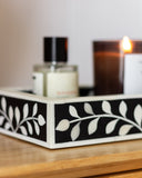Decorative tray with leaf pattern on a wooden surface next to a bottle and a lit candle.