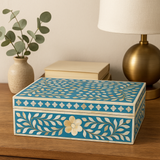 Decorative box with floral patterns on a wooden surface next to a lamp and plant.