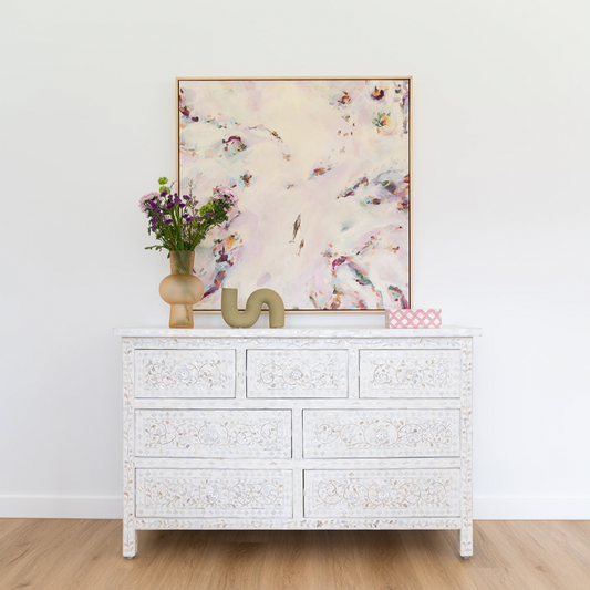 Decorative dresser with a floral painting, vase, and sculpture against a white wall.