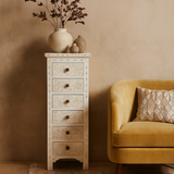 Tall white drawer unit with decorative elements next to a yellow sofa with a patterned pillow against a beige wall.
