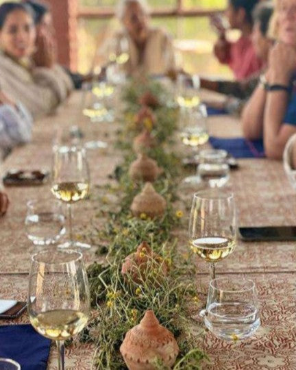 Long table set for a meal with wine glasses and decorative elements, people seated around.