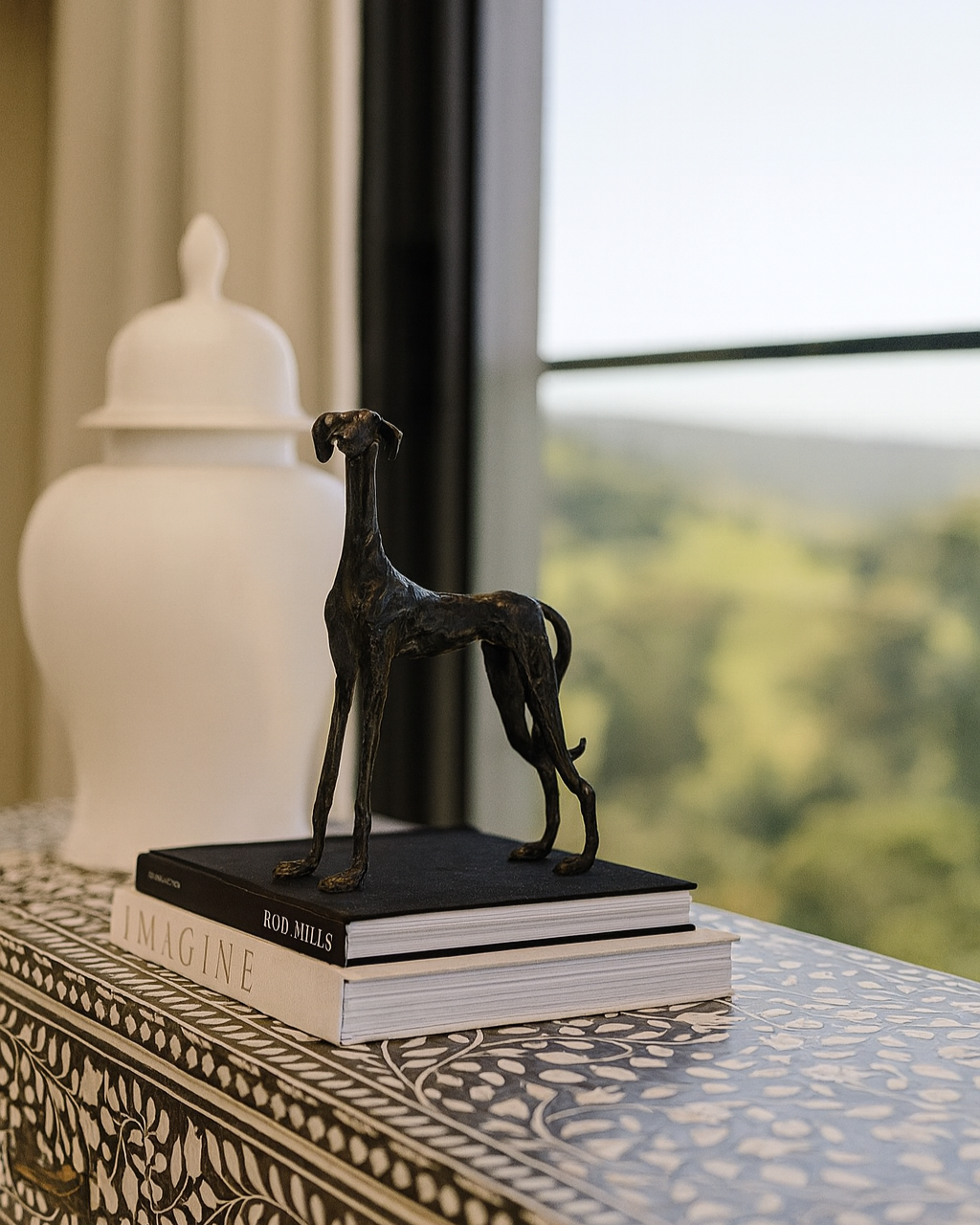 Bronze dog sculpture on books with a white vase and blurred outdoor view in the background