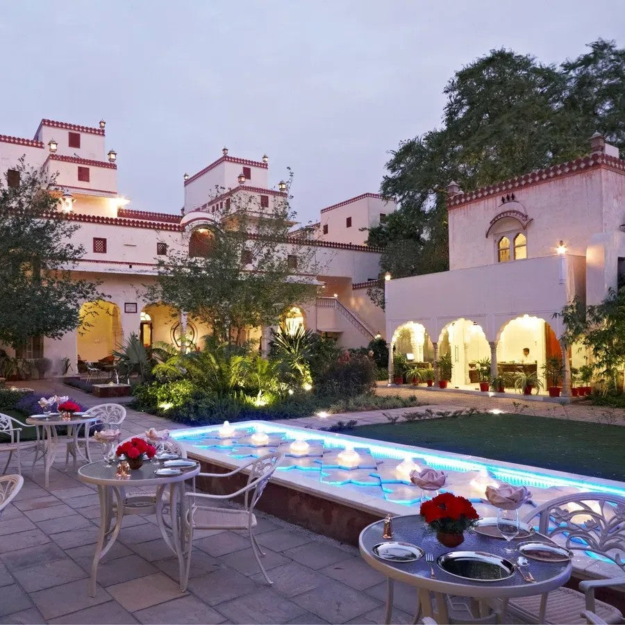 Evening outdoor setting with a pool, tables, chairs, and decorative elements in front of a building.