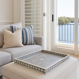 Gray sofa with striped and beige pillows, decorative tray on ottoman, and view of water through window.