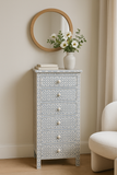 Decorative chest of drawers with a patterned design, round mirror, and vase with flowers on a beige wall.