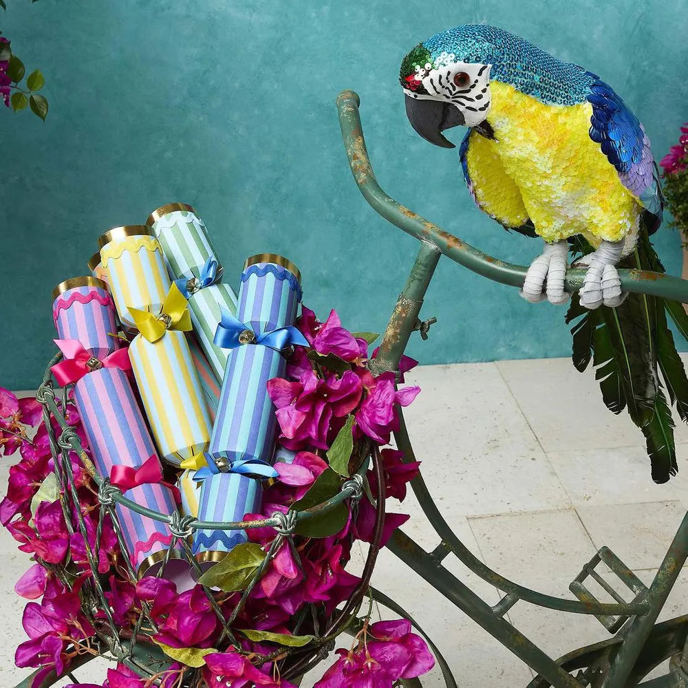 Parrot perched on a bicycle with colorful cracker decorations and pink flowers against a teal wall.