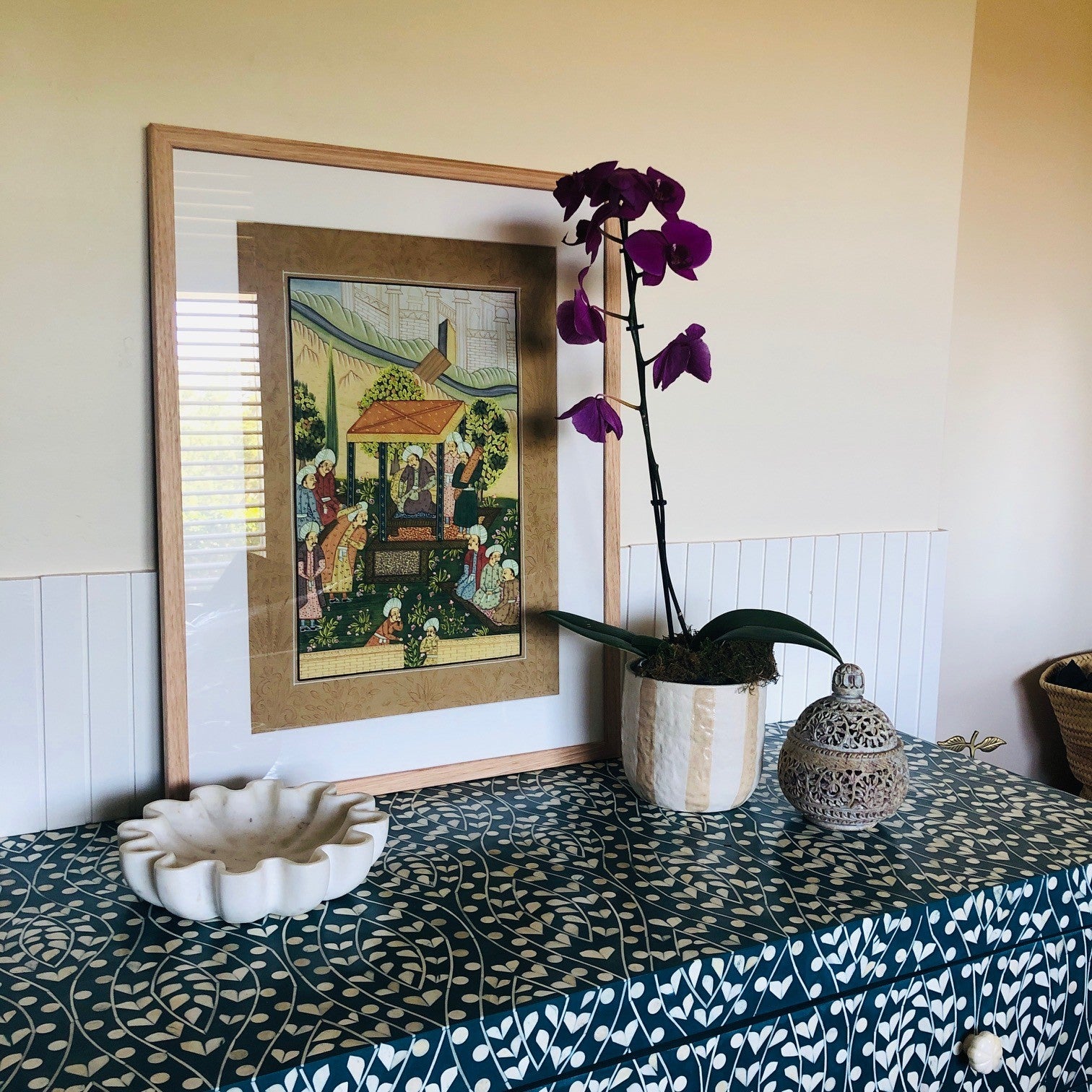 Framed artwork on a table with a purple orchid and decorative items.